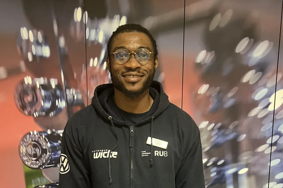 Eine Person mit kurzen Haaren und runder Brille lächelt in die Kamera. Sie trägt einen dunklen Hoodie mit Reißverschluss und steht vor einem Hintergrund mit glänzenden, runden Objekten und unscharfen Lichtreflexen.