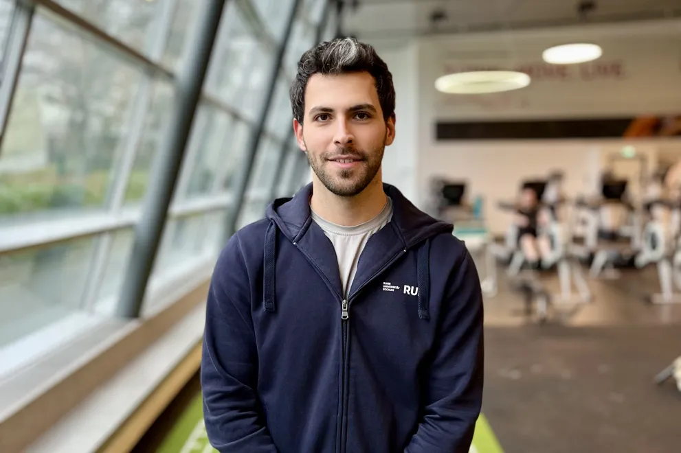 Eine Person mit kurzen dunklen Haaren und Bart steht lächelnd in einem Fitnessstudio und trägt einen marineblauen Zip-Hoodie mit dem Aufdruck der Ruhr-Universität Bochum. Im unscharfen Hintergrund sind Fahrradergometer zu sehen.