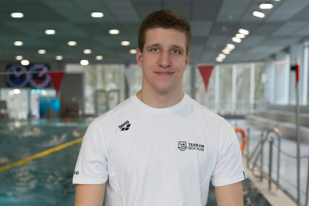 A person is standing in an indoor swimming pool area in front of a lap pool. They are wearing a white athletic T-shirt and looking at the camera with a slight smile. In the background, the water, lane lines, windows, and overhead lights are visible.