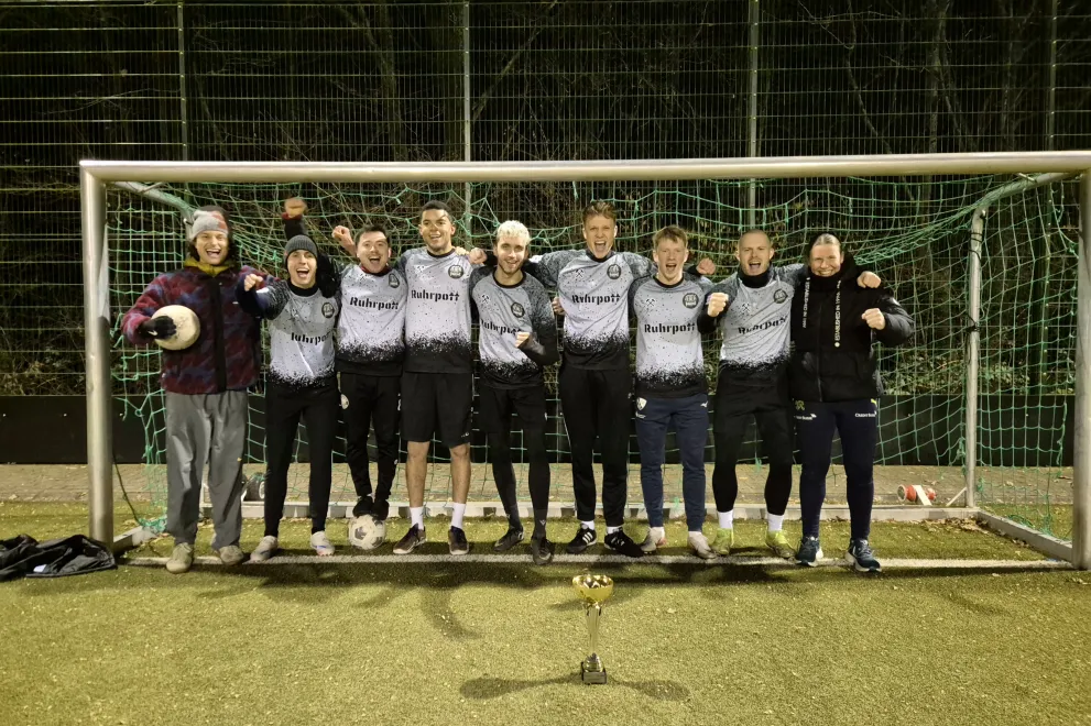 Auf dem Foto ist eine Gruppe von Personen zu sehen, die auf einem Kunstrasenplatz in einem Tor stehen und jubeln.