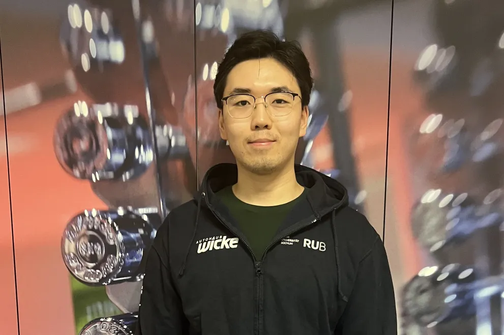 A person stands in a gym in front of a rack of dumbbells. They are wearing a dark hoodie and dark pants, with their arms relaxed at their sides. The background shows rows of weights and is slightly blurred, emphasizing the person in the foreground.