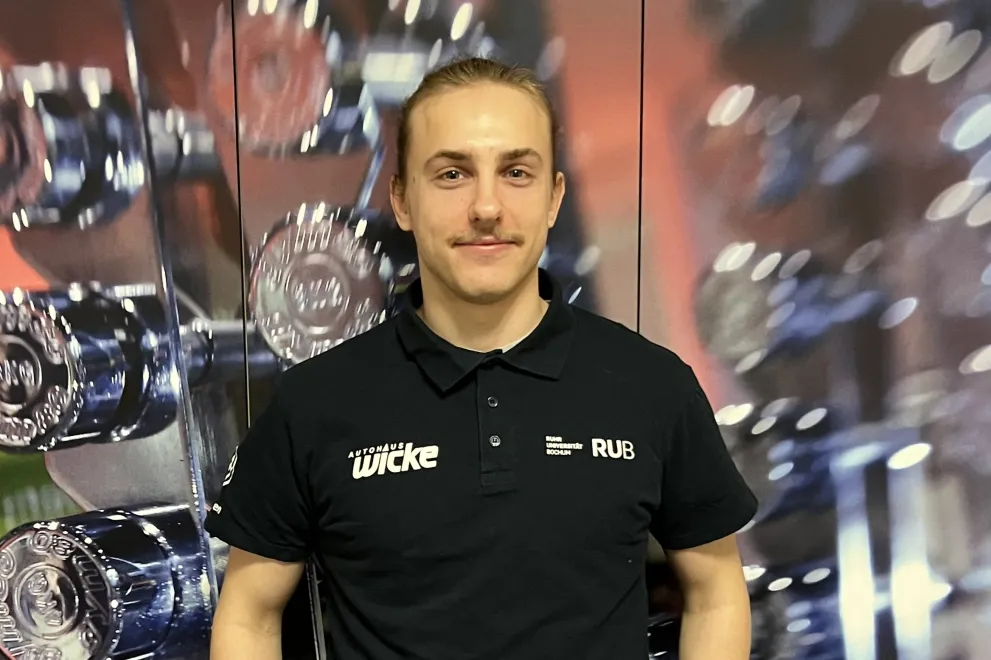 Eine Person steht in einem Fitnessstudio vor einem Hantelregal. Sie trägt ein schwarzes Shirt und eine helle Hose, eine Hand ruht auf der Hüfte. Der Hintergrund ist leicht unscharf, wodurch die Gewichte betont werden und eine räumliche Tiefe entsteht.