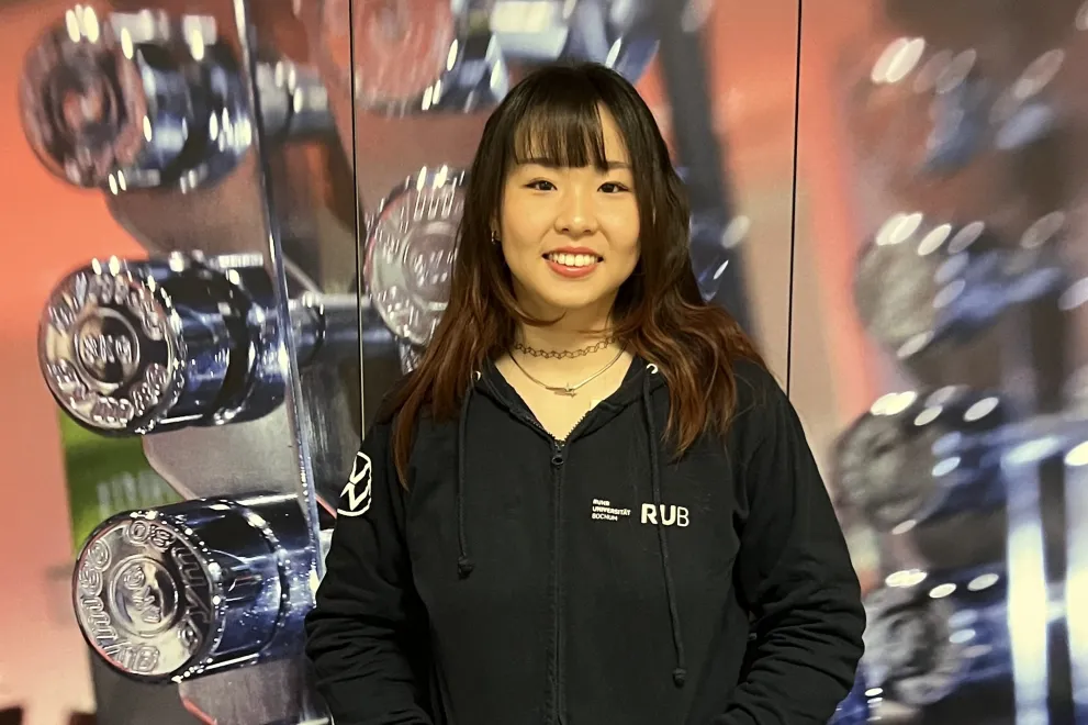 Eine Person steht vor einer Wand mit mehreren glänzenden Hanteln. Sie trägt einen schwarzen Hoodie mit „RUB“-Logo und einen dunklen Rock. Die Hände sind in den Taschen, und sie lächelt in die Kamera. Das Foto wirkt wie in einem Fitnessstudio aufgenommen.