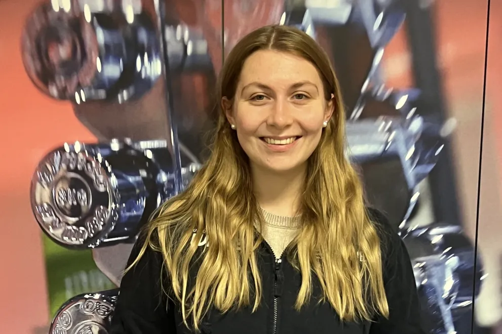 Eine Person steht vor einer Wand mit aufgereihten, glänzenden Hanteln. Sie trägt eine schwarze Zip-Jacke und helle Hose, lächelt in die Kamera und hat die Hände in den Jackentaschen. Das Bild wirkt wie in einem Fitnessstudio aufgenommen.