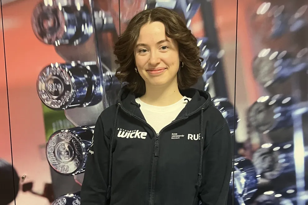 ine Person steht vor einer Wand mit vielen glänzenden Hanteln im Hintergrund. Sie trägt einen schwarzen Hoodie mit Fitnessstudio-Logo und lächelt leicht in die Kamera. Die Hände sind vor dem Körper gefaltet, und das Bild wirkt wie in einem Fitnessstudio oder Trainingsbereich aufgenommen.