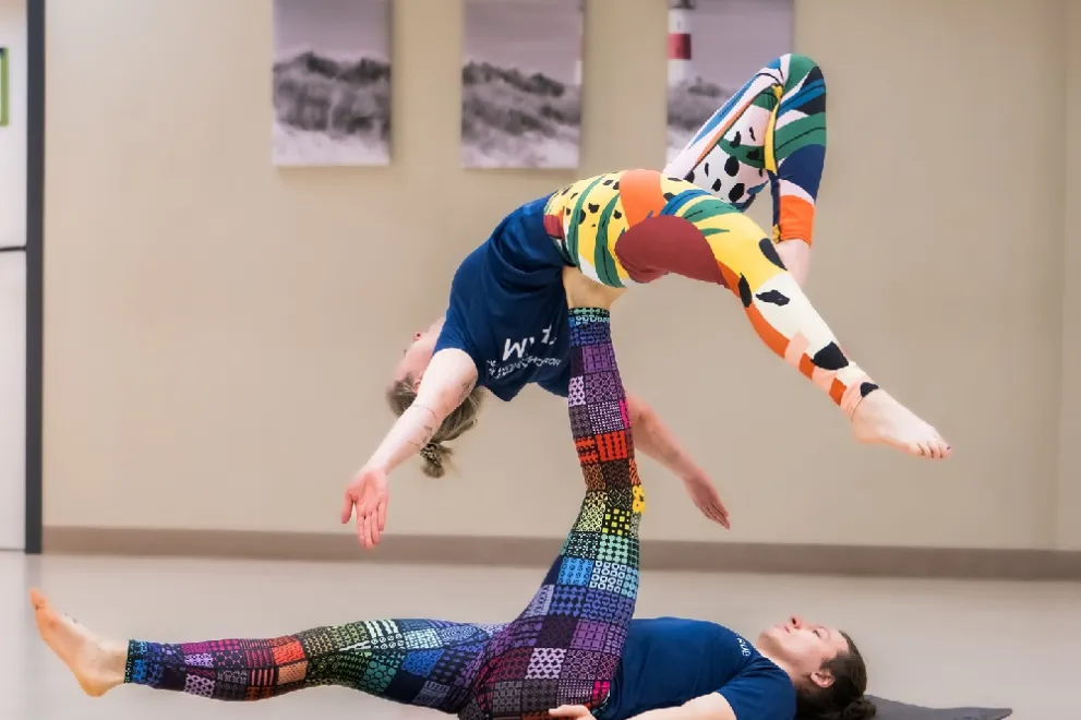 One person lies on the floor with one leg stretched out parallel to the floor and the other leg stretched vertically into the air. Another person lies on the vertical leg with their back arched and both arms stretched out above their head to the side. One of this person's legs falls straight down and the other is bent.