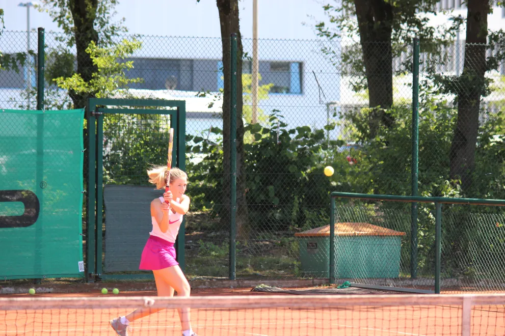A woman is playing tennis on an outdoor court. She is holding her racket up and has the ball in front of her. She takes a step toward the ball.