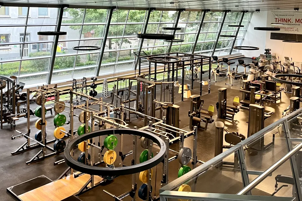 Ein moderner Fitnessbereich in einer großen, lichtdurchfluteten Halle. Zahlreiche Trainingsgeräte wie Hantelbänke, Kraftstationen und Gewichtsscheiben sind ordentlich angeordnet. Große, schräg verlaufende Fensterfronten lassen viel Tageslicht herein und geben den Blick nach draußen frei. Im oberen Bereich verläuft eine Galerie mit Geländer, und an der Wand ist ein motivierender Schriftzug zu sehen. Die Atmosphäre wirkt aufgeräumt und aktiv.