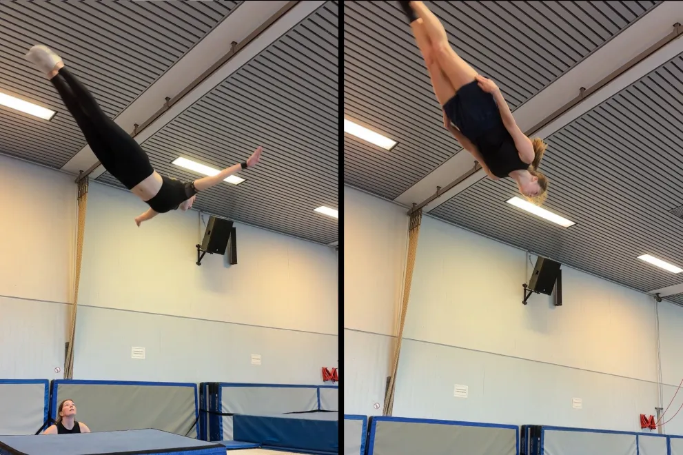 bildbeschreibung 1-2 sätze  Eine Person führt in einer Turnhalle einen Sprung auf einem Trampolin aus und ist hoch in der Luft zu sehen. Unter ihr stehen Sicherheitsmatten, während eine andere Person vom Rand aus nach oben schaut.