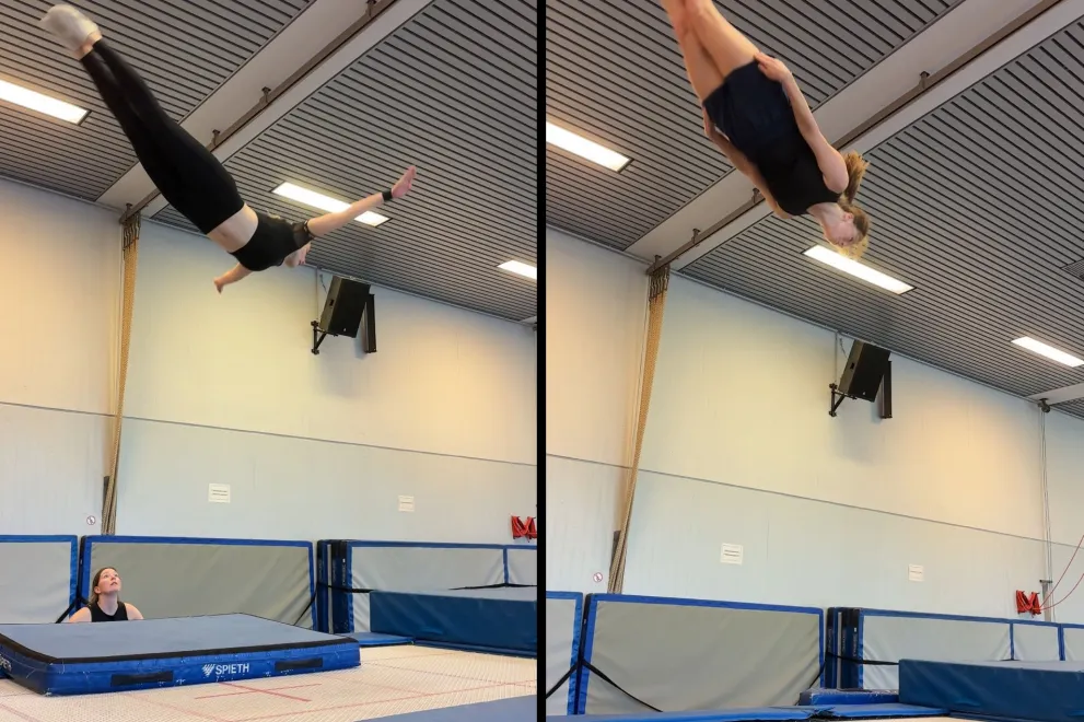  A person is performing a jump on a trampoline in a gym and can be seen high in the air. Safety mats are spread out below them, while another person looks up from the edge.