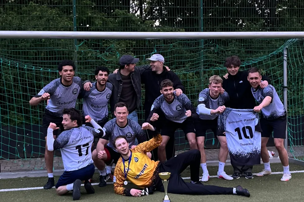 Die Mannschaft von Ruhrpott-PANNA feiert nach dem Finalsieg ausgelassen auf dem Kunstrasenplatz des Hochschulsports Bochum. Mit Pokal, Medaillen und einem Trikot präsentiert sich das Team stolz nach einer perfekten Saison und dem Gewinn der Meisterschaft.
