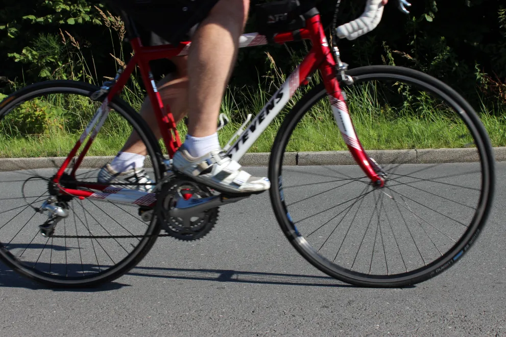 The photo shows a person riding a red racing bike on a paved road. The narrow tires, the hunched riding position, and the curved handlebars are typical features of a racing bike. In the background, there are green spaces and vegetation, suggesting a route through a natural setting.